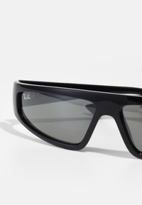 Juodi saulės akiniai su elegantišku dizainu, stačiakampiais objektyvais ir tamsiai pilkų tonų stiklu. Rėmo šone matomas Ray-Ban logotipas.