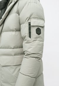 Lys grå polstret vinterjakkeærme med lynlås- og klaplommer samt et sort sekskantet logoemblem på den øverste arm.