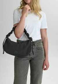 Sort stof håndtaske med en samlet hank, halvmåneform, lynlåslukning og prægede logo detaljer. Båret med en hvid t-shirt og grå jeans.