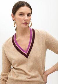 Beżowy sweter dziany z głębokim dekoltem w kształcie V, z fioletowymi i burgundowymi akcentami. Długie rękawy, miękka faktura i dopasowany krój.