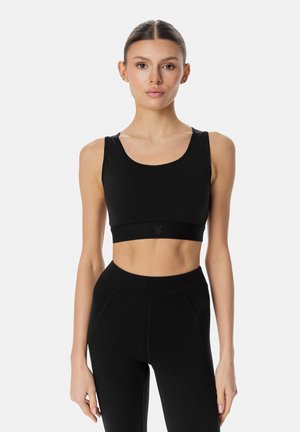 Vrouw met haar naar achter gebonden draagt een zwart mouwloos crop top en leggings met hoge taille tegen een lichte achtergrond.