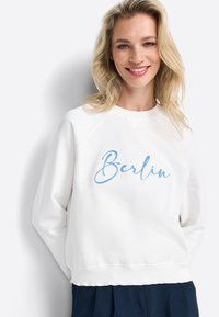 Bijela dukserica s raglan rukavima, s plavim izvezenim natpisom "Berlin". Mekana tekstura i rebraste manžetne, nosi se preko tamne suknje.
