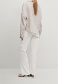 Femme aux cheveux longs blonds portant une veste rayée beige et blanche, un pantalon blanc et des sandales à talons ouvertes beige, debout devant un fond uni.