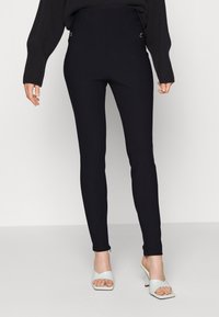 Svarta högmidjade leggings i ett stretchy material. Har sidofickor med guldfärgade detaljer. Matchas med vita öppna klackskor.