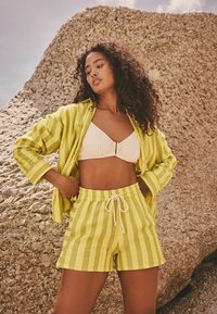 Gestreiftes gelb-weißes Shirt und passende Shorts mit einem Kordelzug-Bund sowie einem strukturierten weißen Bikini-Oberteil. Strukturierten, felsigen Hintergrund.