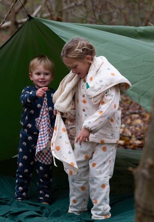 Zwei Kinder in gemusterten Schlafanzügen stehen unter einem grünen Zelt. Das Outfit des Mädchens ist cremefarben mit einem orangefarbenen Sonnen- und blauen Muster; das des Jungen ist marineblau mit Wolken.