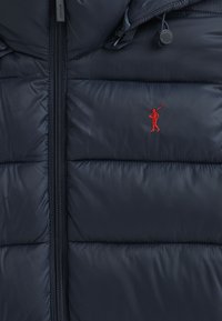 Navy puffersock med quiltad textur, dragkedja framtill, justerbar huva och röd broderad logotyp. Slät, vattenavvisande material.