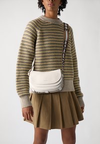 Borsa a tracolla bianca in pelle con forma arrotondata, superficie testurizzata e tracolla regolabile con accento del marchio. Indossata sopra un maglione a righe.