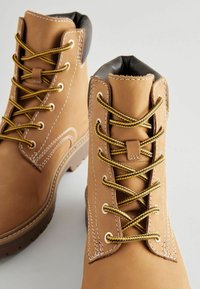 Botas de cuero marrón claro con collar acolchado negro, cordones amarillos y costuras blancas. Suela de goma duradera con patrón de agarre texturizado.