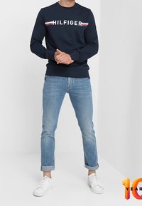 Sudadera azul marino con la marca "HILFIGER" en blanco, acentuada con rayas rojas y blancas; combinada con jeans azul claro y zapatillas blancas.