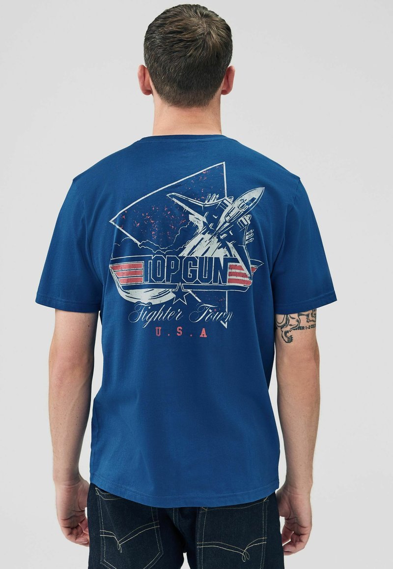 T-shirt in cotone blu con un design grafico di un jet e la scritta "TOP GUN Fighter Town U.S.A." sulla parte posteriore. Vestibilità classica con maniche corte.