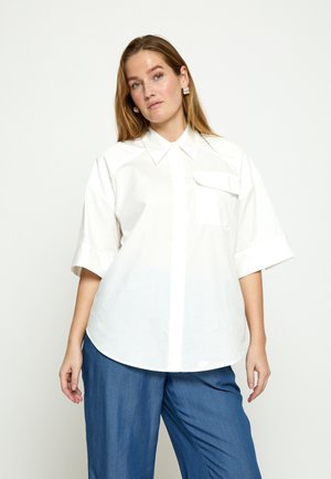 Frau mit langen Haaren, die ein lockeres weißes kurzärmliges Hemd mit Knöpfen und weite blaue Hosen vor einem schlichten Hintergrund trägt.