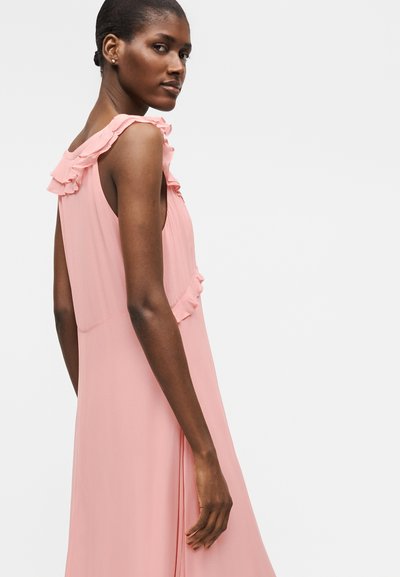 Γυναίκα με σκούρο δέρμα που φοράει ροζ φόρεμα χωρίς μανίκια με βολάν, κοιτάζοντας πάνω από τον ώμο της μπροστά σε απλό ανοιχτό φόντο.