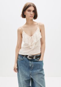 Ung kvinna klädd i en krämfärgad blus med volanger utan ärmar och lösa blå jeans med ett svart bälte, som står framför en vit bakgrund.