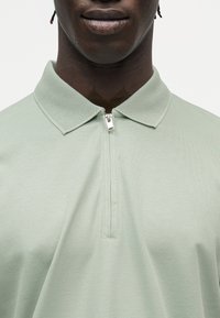 Κοντινό πλάνο ατόμου που φοράει ανοιχτό πράσινο πόλο με φερμουάρ και υφασμένο γιακά, σε λευκό φόντο.