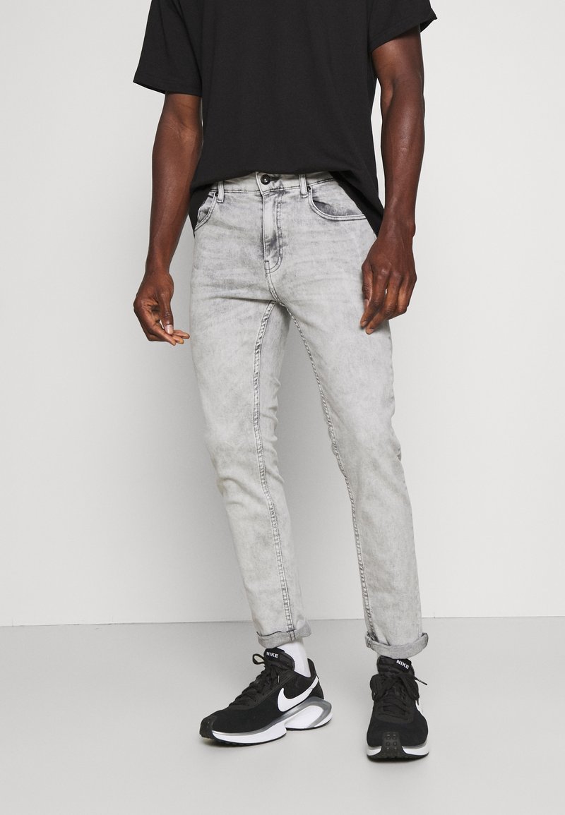 Homme portant un jean gris clair retroussé, un t-shirt noir, des chaussettes blanches et des baskets Nike noires, debout contre un fond clair uni.