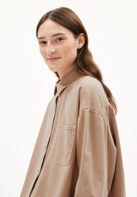 Overmaatse blouse in lichtbruin, met een borstzak, zachte stof en een gekraagde halslijn met zichtbare stikseldetails.