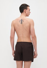 Shorts de bain marron avec ceinture elastique, portés sur un torse nu, montrant un tatouage de fleur de lys sur le haut du dos.