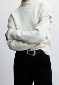 Pull en grosse maille crème avec un col montant, des manches côtelées et une coupe décontractée, associé à un pantalon en velours côtelé noir et une ceinture noire cloutée.