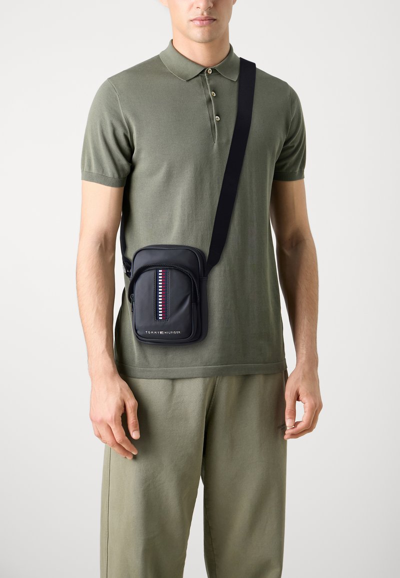 Bolso cruzado azul marino con un bolsillo frontal con cremallera, que presenta un patrón de rayas verticales en rojo, blanco y azul. Material suave y correa ajustable.