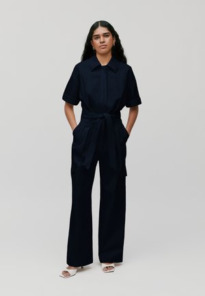 Combinaison bleu marine avec un col rabattu, des manches courtes, une taille cintrée et un pantalon large ; elle dispose de poches et d'une texture lisse.