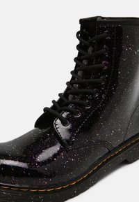Boots noires à lacets avec finition brillante et paillettes multicolores, avec des coutures jaunes sur le bord de la semelle.