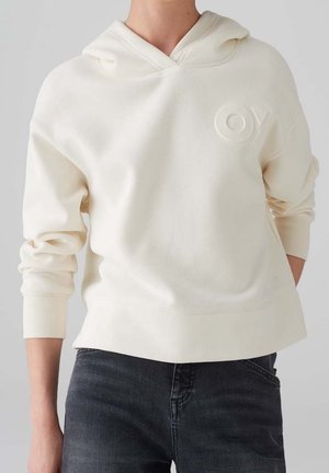 Personne portant un sweat à capuche de couleur crème avec "OY" en relief sur la poitrine et un jean foncé, debout devant un fond clair uni.