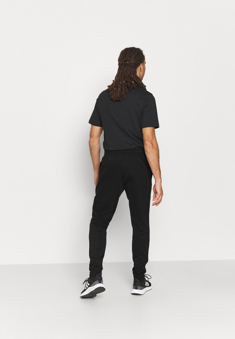 Schwarzes T-Shirt mit kurzen Ärmeln, kombiniert mit schwarzen Jogginghosen. Das Outfit hat einen einfachen, lockeren Schnitt und wird durch schwarze Sportschuhe ergänzt.
