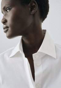 Witte katoenen shirt met een gestructureerde kraag en knoopsluiting, die een gladde textuur en een getailleerde pasvorm laat zien. Zijkant van de kraag en halslijn.