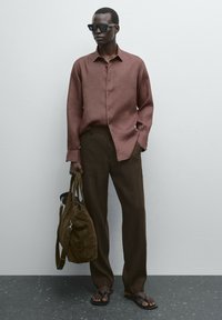 Braune Leinenhemd, lockere braune Hose, schwarze Sonnenbrille und braune Sandalen. Hält eine braune Stofftasche mit einem Schultergurt.