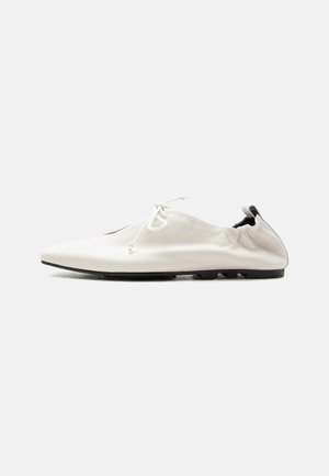 Chaussure blanche minimaliste en cuir à enfiler avec talon rentré, fermeture à lacets et semelle fine noire sur fond uni.