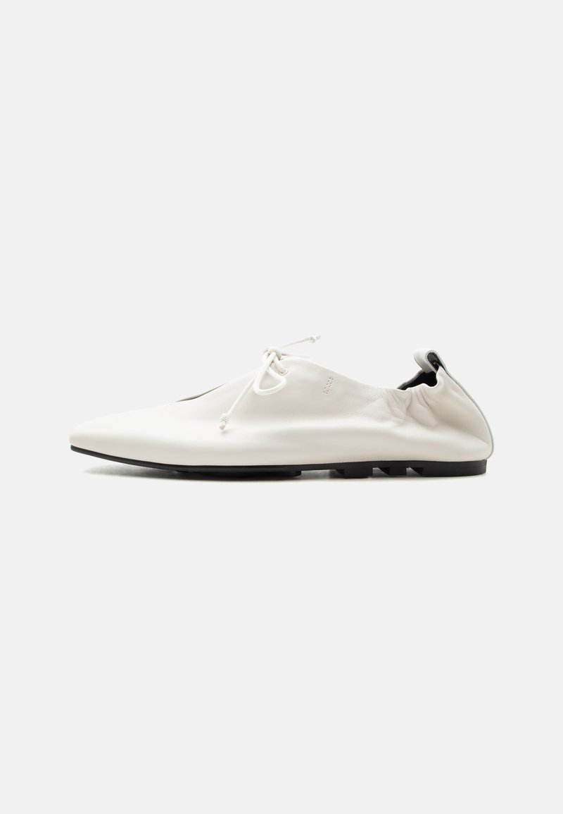 Chaussure blanche minimaliste en cuir à enfiler avec talon rentré, fermeture à lacets et semelle fine noire sur fond uni.