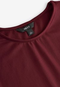 Burgundská, krátkosrstá blúzka s okrúhlym výstrihom a zberovými detailmi na prednej strane. Tkanina vyzerá hladko a mäkko. Viditeľná nálepka s logom značky.