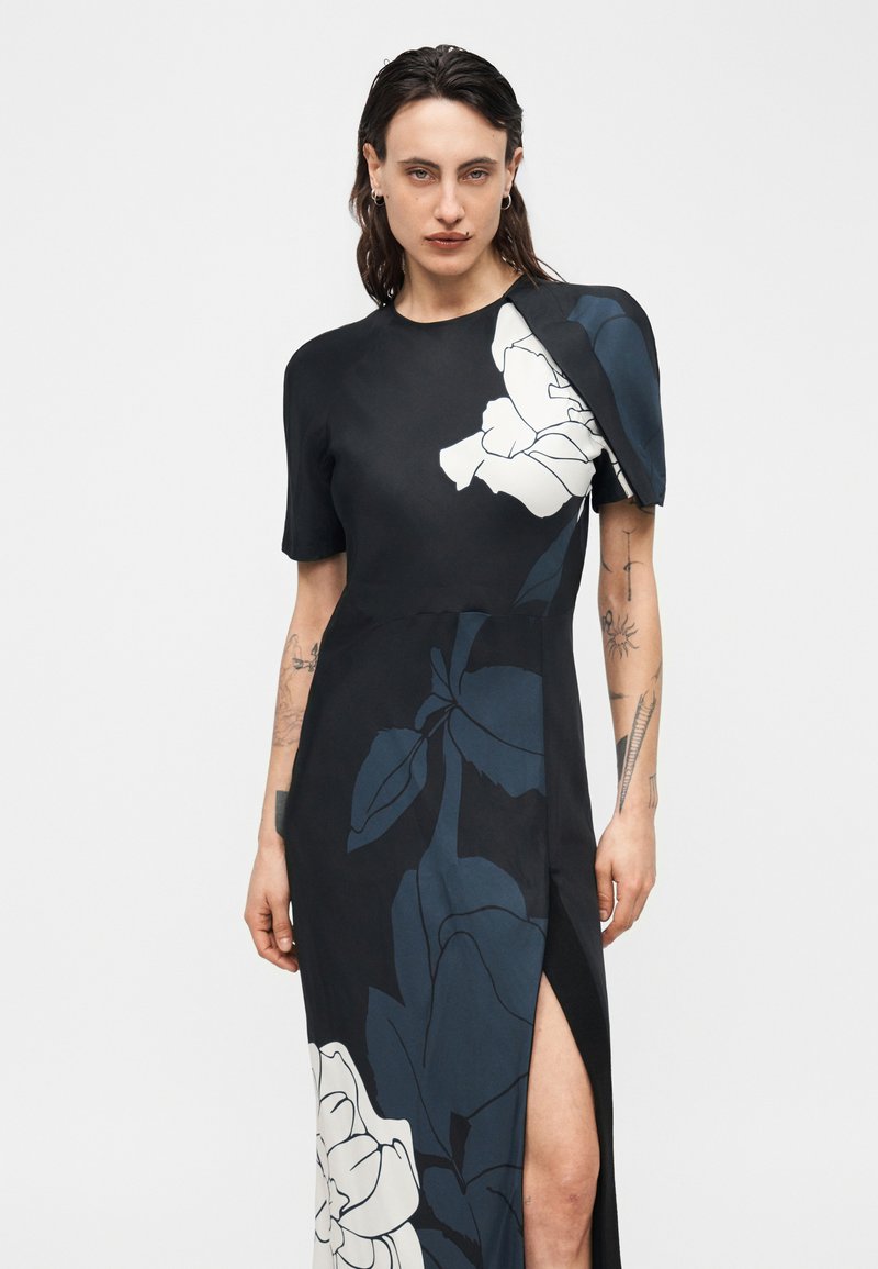 Femme portant une robe noire à grands motifs floraux bleus et blancs, manches courtes, détails aux épaules, avec une fente haute sur la cuisse.