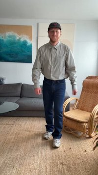 Indivíduo com uma camisa xadrez e jeans escuros, usando ténis cinzentos e um boné preto, a caminhar numa sala de estar clara e minimalista.