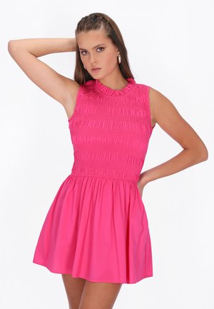 Kobieta w różowej, bezrękawnej sukience z fakturowanym gorsetem i rozkloszowanym dołem, pozująca z jedną ręką założoną za głowę, a drugą na biodrze.