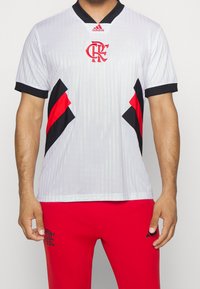 Maillot de sport blanc avec des rayures verticales, col noir, accents rouges et logo. Associé à un pantalon rouge avec un logo noir.