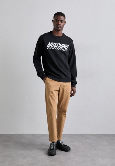 Sweat-shirt noir avec "Moschino Couture!" en texte blanc, associé à un pantalon beige et des chaussures à plateforme noires avec des lacets argentés.