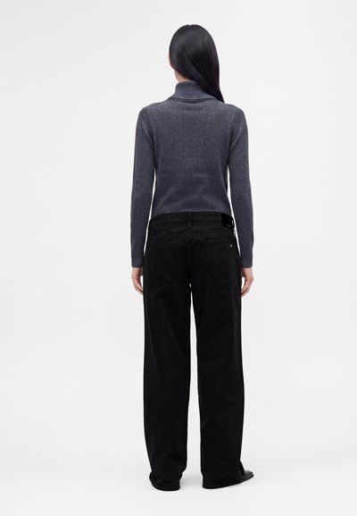 Femme aux longs cheveux foncés portant un pull gris à col roulé et un jean noir ample, debout de dos sur un fond blanc.