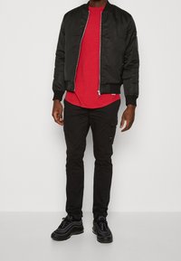 Černá bombera přes červené tričko, doplněná o černé nákladní kalhoty a černé tenisky s vrstveným designem.