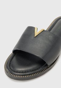 Sandale glissante en cuir noir avec une large sangle, mettant en avant un accent doré en forme de V. La semelle a une finition noire texturée avec des détails de couture dorés.