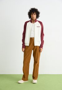White and maroon zip-up jacket with logo, white tank top, brown corduroy pants, and light sneakers on green flooring.