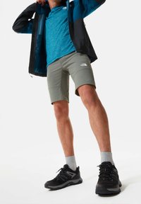 Personne portant une veste noire et bleue, une chemise bleue, un short gris, des chaussettes grises et des chaussures de randonnée noires se tenant sur un fond blanc.