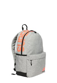 Mochila de tela gris con un gran logotipo naranja "SUPER", acentos negros, un bolsillo frontal y cierre de cremallera. Diseño ergonómico y correas ajustables.