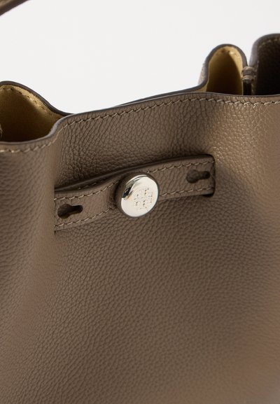 Közelkép egy taupe színű, texturált bőrtáskáról, ezüst gombbal, amelyen gravírozott logó található, és részletes varrás a felső szélen.