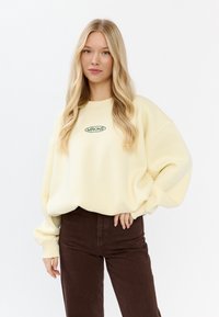 Jasnożółty, oversize'owy sweter z okrągłym, zielonym logo na piersi, w połączeniu z ciemnobrązowymi spodniami z kordurowej tkaniny. Miękki, casualowy materiał.