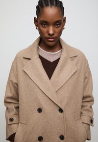 Beige double-breasted coat in a textured wool blend, featuring large black buttons, wide lapels, and a relaxed fit, layered over a brown sweater.