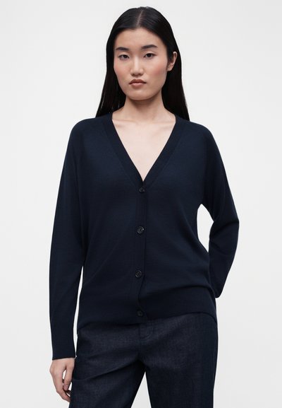 Νέα γυναίκα με μακριά μαύρα μαλλιά, ντυμένη με μπλε σκούρο πλεκτό ζακέτα με κουμπιά και σκούρο παντελόνι, στέκεται με το ένα χέρι πίσω από την πλάτη της.