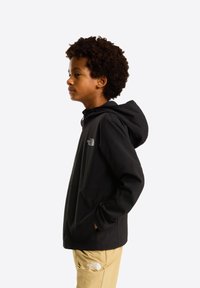 Jongen in een zwart jack met capuchon en beige broek met The North Face-logo's, staand in profiel met handen in de zakken tegen een witte achtergrond.