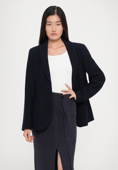 Ženska nosi temno modri blazer preko bele majice in temno modri krilu z odprtino spredaj ter s pasom na zavozlji, stoji središče z eno roko na boku.